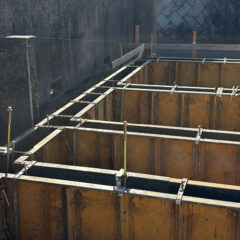 Concrete formwork in a rectangular pit, supported by metal frames and scaffolding with vertical rods protruding upward at a construction site