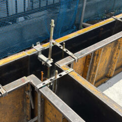 Construction site with steel formwork panels, clamps, and a vertical rebar; blue safety netting in the background.