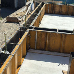 Construction site showing wooden formwork and metal braces around a concrete foundation slab scaffolded for pouring.