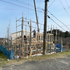 Wood-framed house under construction with scaffolding and workers on the frame near a utility pole.