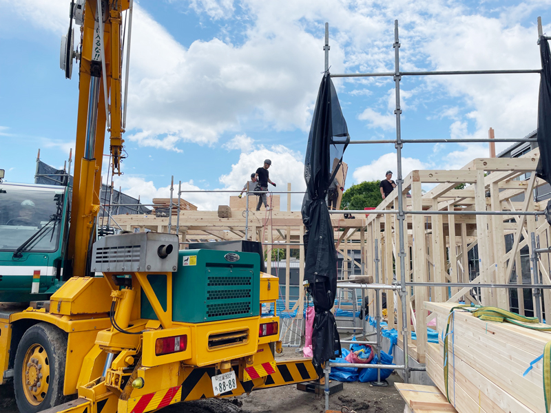 Construction site with a yellow crane truck and wooden framing, workers on the scaffolded upper level under a blue sky.