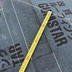 Yellow tape measure laid diagonally across dark construction underlayment panels with printed markings.