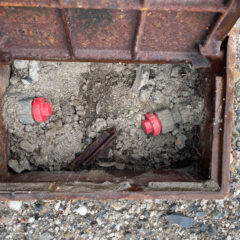 Open rusty manhole with two red pipe caps surrounded by dirt and rubble on a gravel surface, showing interior pipes.
