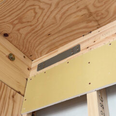 Close-up of wooden framing with a metal connector plate securing two boards at a corner during construction, with yellow drywall sheathing nearby.