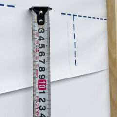 Tape measure held against a white surface with blue dashed marks, showing about 10 cm; a wooden post on the right.