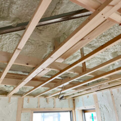 Unfinished ceiling with wooden framing, spray foam insulation, and exposed pipes in a room under construction.