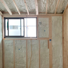 Interior of a partially insulated room under construction with exposed wooden studs, spray foam insulation, and a small window; an orange cord hangs from a wall outlet nearby.
