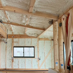 Interior of a house under construction with exposed wooden studs, spray foam insulation on walls and ceiling, and a small window in the wall.