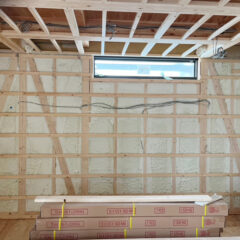 Construction room with exposed wooden framing, insulation, a small horizontal window, and stacked flooring boxes in the foreground.