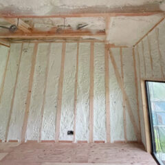 Unfinished interior scene with spray foam insulation on walls, exposed wooden studs, plumbing/electrical lines, and a glass door to the outside.