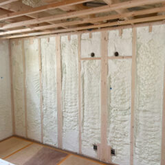 Construction site interior with exposed wooden studs and sprayed foam insulation between them; ceiling framing and loose wires visible.