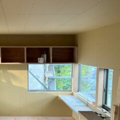 Room under construction with beige drywall, open upper cabinets, and a window seat area; construction materials visible on the shelves outside.