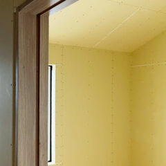 Interior of a room under construction with yellow drywall panels, a wooden door frame, and a narrow vertical window on the left side.