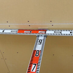 Construction tape measure stretched horizontally across a wall, with a second tape measure running vertically downward, showing numbers like 7, 8, 9; used to measure and mark dimensions on drywall panels.