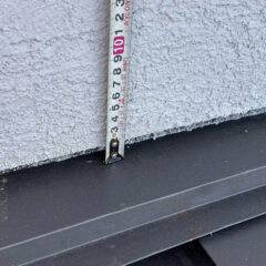 Measuring tape extended vertically along a textured gray exterior wall, showing about 10 cm from the ground to the bottom edge of a metal surface.