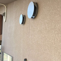 Exterior beige stucco wall featuring two white circular wall vents; scaffolding and construction materials visible on the left side.