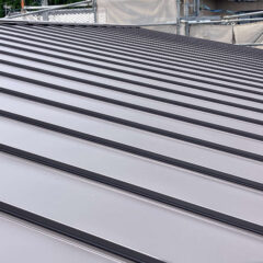 Gray standing-seam metal roof panels on a sloped roof, with scaffolding in the background indicating construction work.