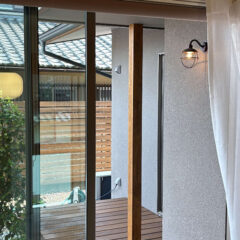 Interior view of a sliding glass door opening to a wooden deck, with a vertical wooden post and sheer white curtain on the right.