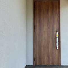 Wooden front door set in light gray stucco walls; a tall white vertical strap-like object is mounted to the right side of the door.