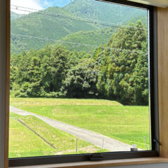 View through a large window of a green field, a gravel path, and a forested hillside beyond.