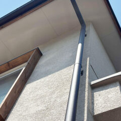 Low-angle view of a house corner with a gray downspout along a textured white wall, a wooden-framed window, and an overhanging eave.