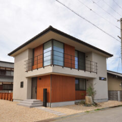 Two-story house with wood-colored panels, beige exterior, and a metal balcony on the upper floor, on a quiet street with gravel yard.