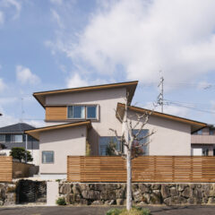 Modern beige, multi‑level house with a slanted roof behind a wooden fence and a bare tree in the foreground. a stone retaining wall along the street.