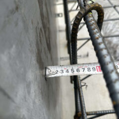 Close-up of a tape measure stretched along a rough concrete wall, with rusted rebar bars in the foreground at a construction site.