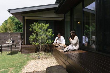 A man and woman sit on a wooden deck with two small dogs, taking photos outside a modern glass-walled house.
