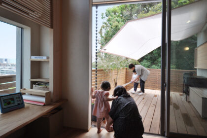 Interior room with a sliding glass door opening to a wooden deck; two small children and an adult near the door, while another person on the deck tends a white canopy shade.