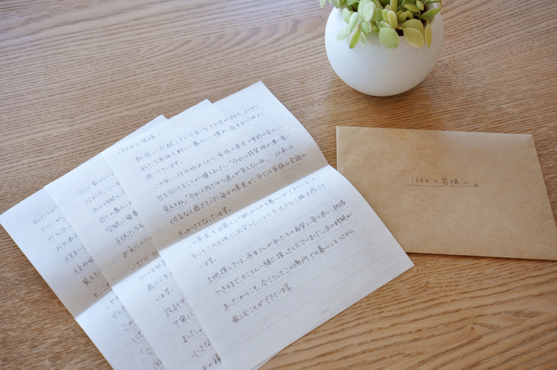 Open handwritten letters in Japanese on a light wooden table, next to a brown envelope and a small potted plant.