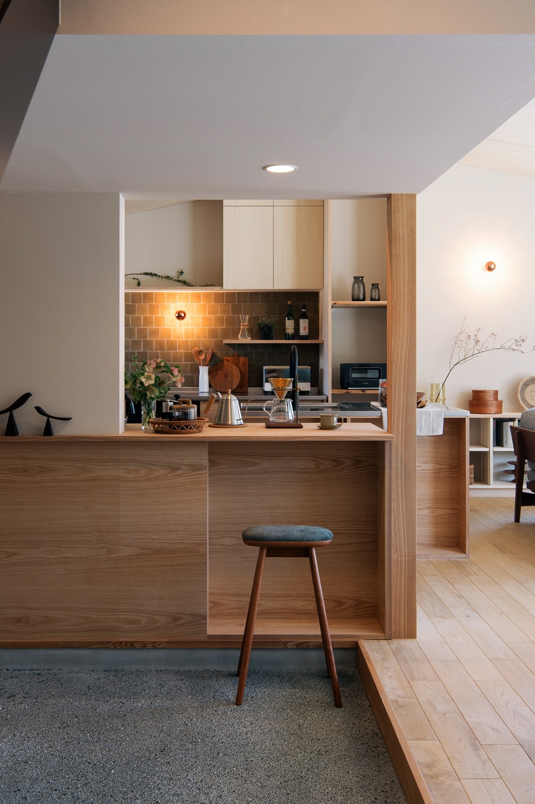 Open wooden kitchen with a bar counter, teal stool, and a tiled backsplash; plants and coffee gear on the counter.