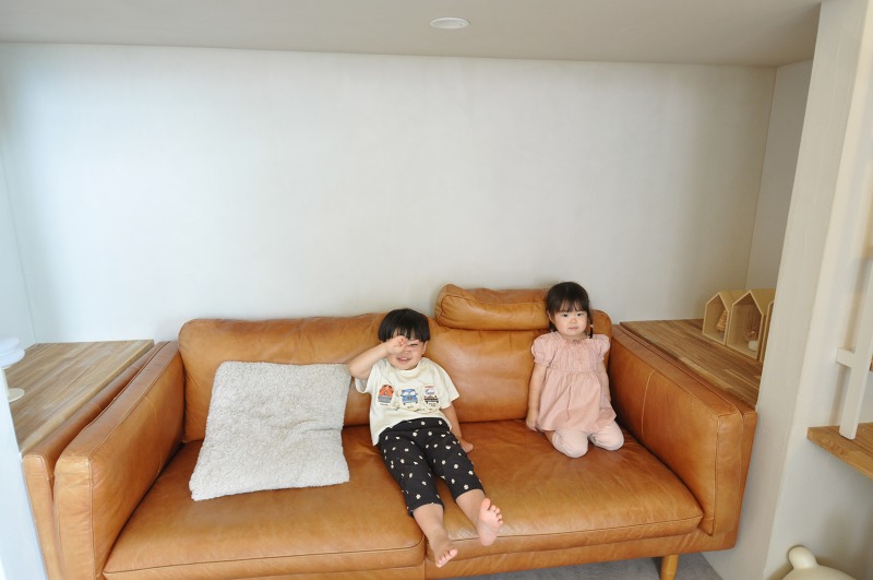 Two children sit on a tan leather sofa in a bright living room; one waves while the other sits in a pink dress.